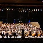 A symphony orchestra sits in chairs on a stage, prepared to begin playing. Between the musicians and the darkened auditorium, conductor raises a left hand. Members of the chorus stand on risers behind the orchestra, backed by an acoustical shell beneath dozens of floodlights.