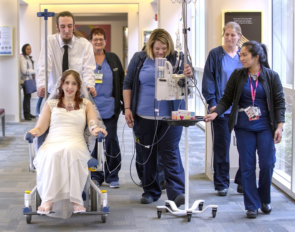A man in a white shirt and black tie pushes a woman in a wedding gown in a wheelchair down a hospital corridor. With him are several members of the hospital staff in blue nurse’s uniforms. The nurse in the center wheels a tall stand from which dangle wires which are attached to the woman in the gown.