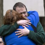 Bill Griffis’ eyes are closed behind his glasses as he embraces Brooke Olenowski, whose back is facing the camera. They are bracketed by two trees in the background, where another woman looks on.