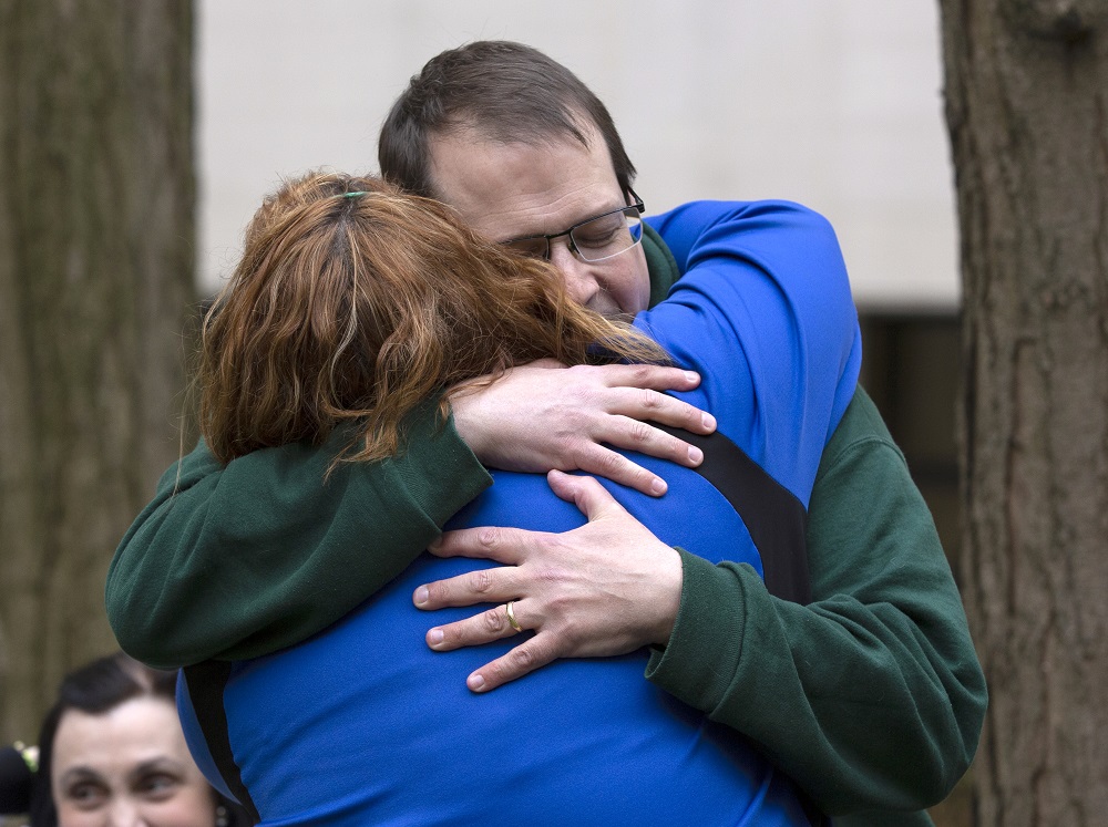 Bill Griffis’ eyes are closed behind his glasses as he embraces Brooke Olenowski, whose back is facing the camera. They are bracketed by two trees in the background, where another woman looks on.