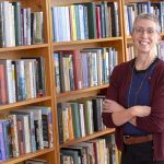 Bernice Hausman, chair of Penn State College of Medicine’s Department of Humanities, stands in a library with her arms crossed and smiling. She has short, gray hair and is wearing glasses, a sweater, a T-shirt, casual pants and a brown belt. Five rows of bookshelves filled with books are to her left.