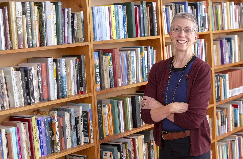 Bernice Hausman, chair of Penn State College of Medicine’s Department of Humanities, stands in a library with her arms crossed and smiling. She has short, gray hair and is wearing glasses, a sweater, a T-shirt, casual pants and a brown belt. Five rows of bookshelves filled with books are to her left.