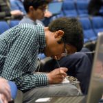 Cedar Cliff High School junior Thussentham Walter-Angelo, winner of the 2019 Central PA Regional Brain Bee, bends over a clipboard on his lap and writes answers during the 2019 USA National Brain Bee, held April 12-14 at Penn State College of Medicine. He is seated in an auditorium, surrounded by other students. He wears a plaid shirt, gray pants and glasses.