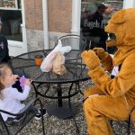 A person dressed as the Penn State Nittany Lion sits at an outdoor table with a little girl and a man in a jacket and a ball cap. They are reflected in the window of a shop advertising bagels.