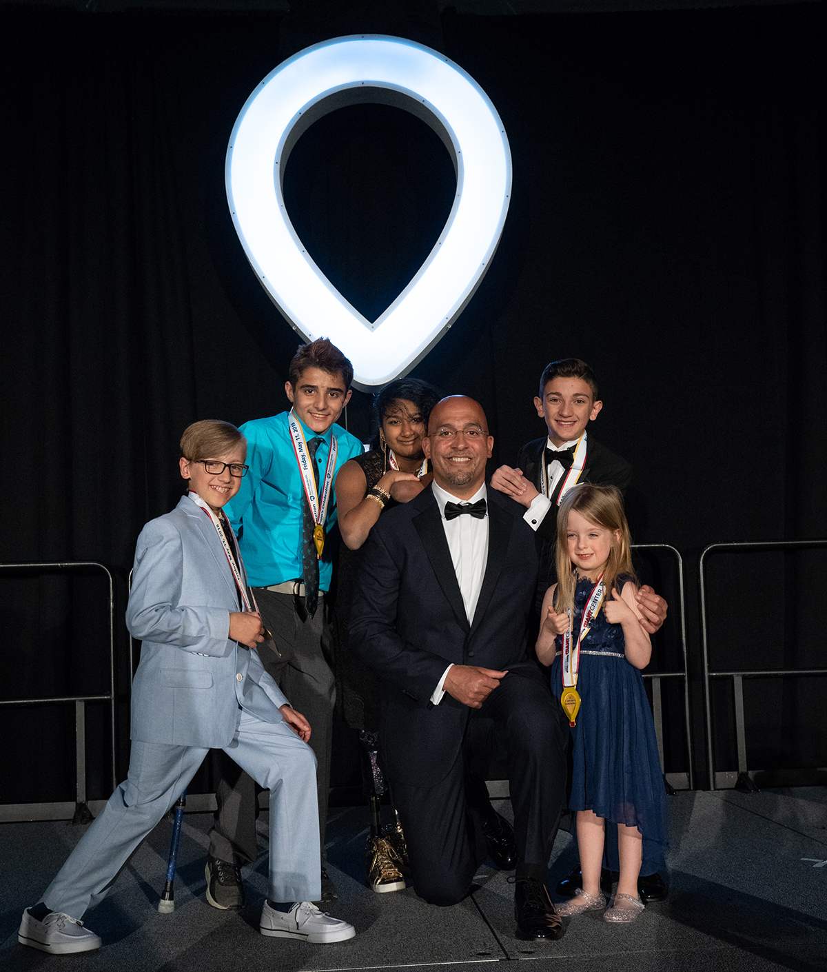 Penn State Football Coach James Franklin, wearing a tuxedo, kneels on one knee. Around him are five young children. An illuminated CMN logo is in the background.