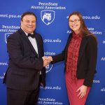 A woman in a dress and blazer shakes hands with a man in a suit with a bow tie. Behind them is a surface covered in the logo and web address for the Penn State Alumni Association.