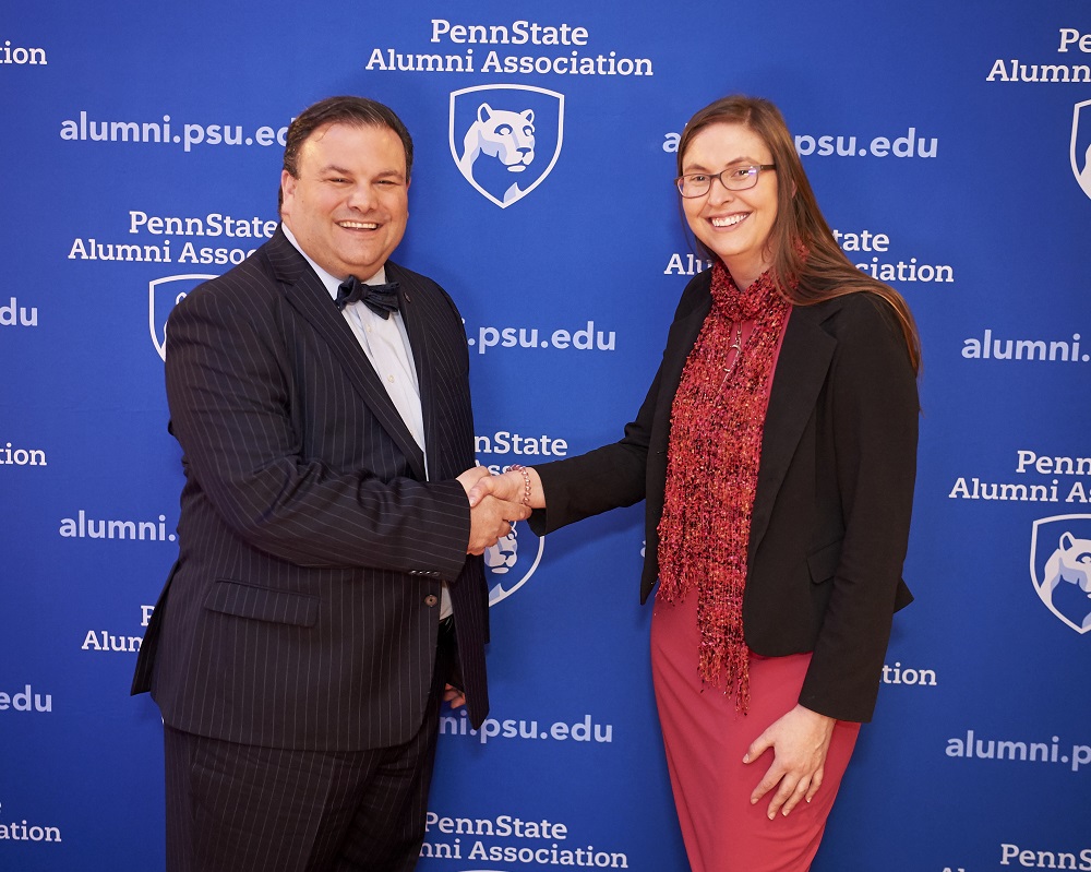 A woman in a dress and blazer shakes hands with a man in a suit with a bow tie. Behind them is a surface covered in the logo and web address for the Penn State Alumni Association.