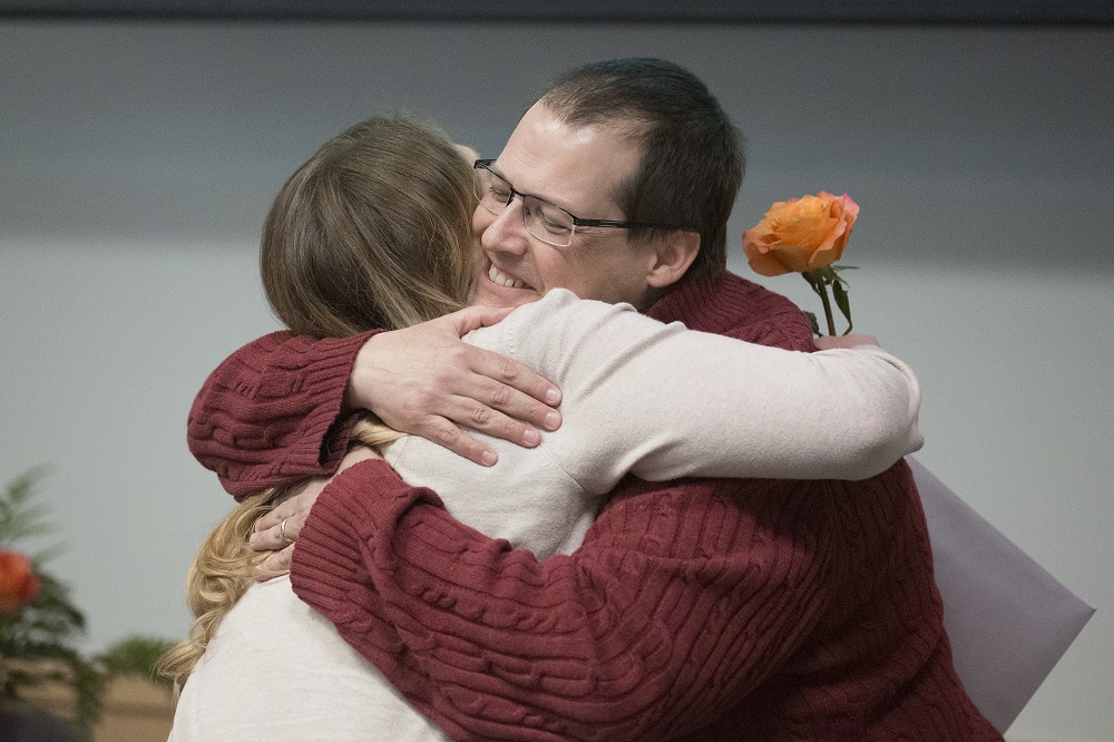 A man with glasses smiles as he hugs a woman. The woman hand on the man’s shoulder blade holds a rose.