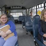 Penn State Health employees Marion Zakrzewski, left, and Cathy Laguna ride in the Penn State Health shuttle. Zakrzewski is wearing a sweater and pants and holding a spiral-bound notebook. Laguna is wearing a dress and a vest and has a spiral-bound notebook on her lap. A man sits in the back of the shuttle. Cars are visible through windows on both sides of the shuttle.