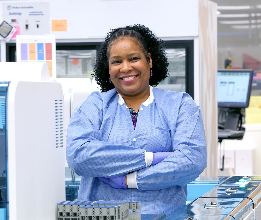 Senior technical specialist Terri Neibauer stands in front of a conveyor belt in the Automated Testing Lab at Penn State Health Milton S. Hershey Medical Center. She has short, curly hair and is wearing a lab smock and medical gloves. The conveyor belt has sections of plastic covers on top. Behind her are lab testing machines and a computer.