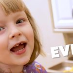 A little girl is saying something in a room with a cabinet and a sink. Her name “EVEY” is written on a slant in block type in the foreground.