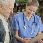 A woman in scrubs with a stethoscope draped over her neck smiles as she looks at a clipboard with a man in a sweater. Both sit on a leather couch. Behind them, house plants sit on shelves on wood-paneled walls.