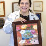 Dr. Zakiyah Kadry of Hershey Medical Center holds an oil painting created for her by a patient. She is wearing a white lab coat, top and gold necklace. The painting is in a wooden frame and shows flowers, Don Quixote, a coffee cup and a thank you note. Behind her are diplomas and plaques.