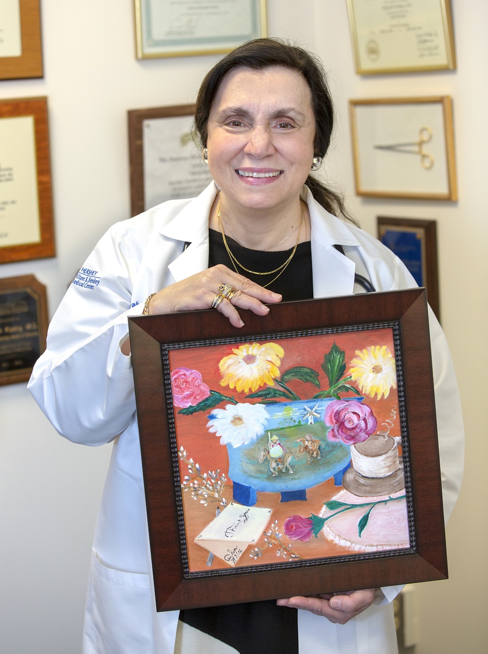 Dr. Zakiyah Kadry of Hershey Medical Center holds an oil painting created for her by a patient. She is wearing a white lab coat, top and gold necklace. The painting is in a wooden frame and shows flowers, Don Quixote, a coffee cup and a thank you note. Behind her are diplomas and plaques.