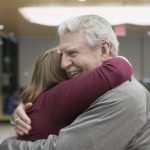 Ed Frederick hugs Dr. Elizabeth Werley at the first “See Me Now” program. Ed has white hair and a moustache and is wearing a gray sweatshirt. Dr. Werley is wearing a long-sleeved sweater. Behind them are cafeteria tables and chairs. A large light fixture is above them.