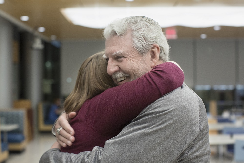 Ed Frederick hugs Dr. Elizabeth Werley at the first “See Me Now” program. Ed has white hair and a moustache and is wearing a gray sweatshirt. Dr. Werley is wearing a long-sleeved sweater. Behind them are cafeteria tables and chairs. A large light fixture is above them.