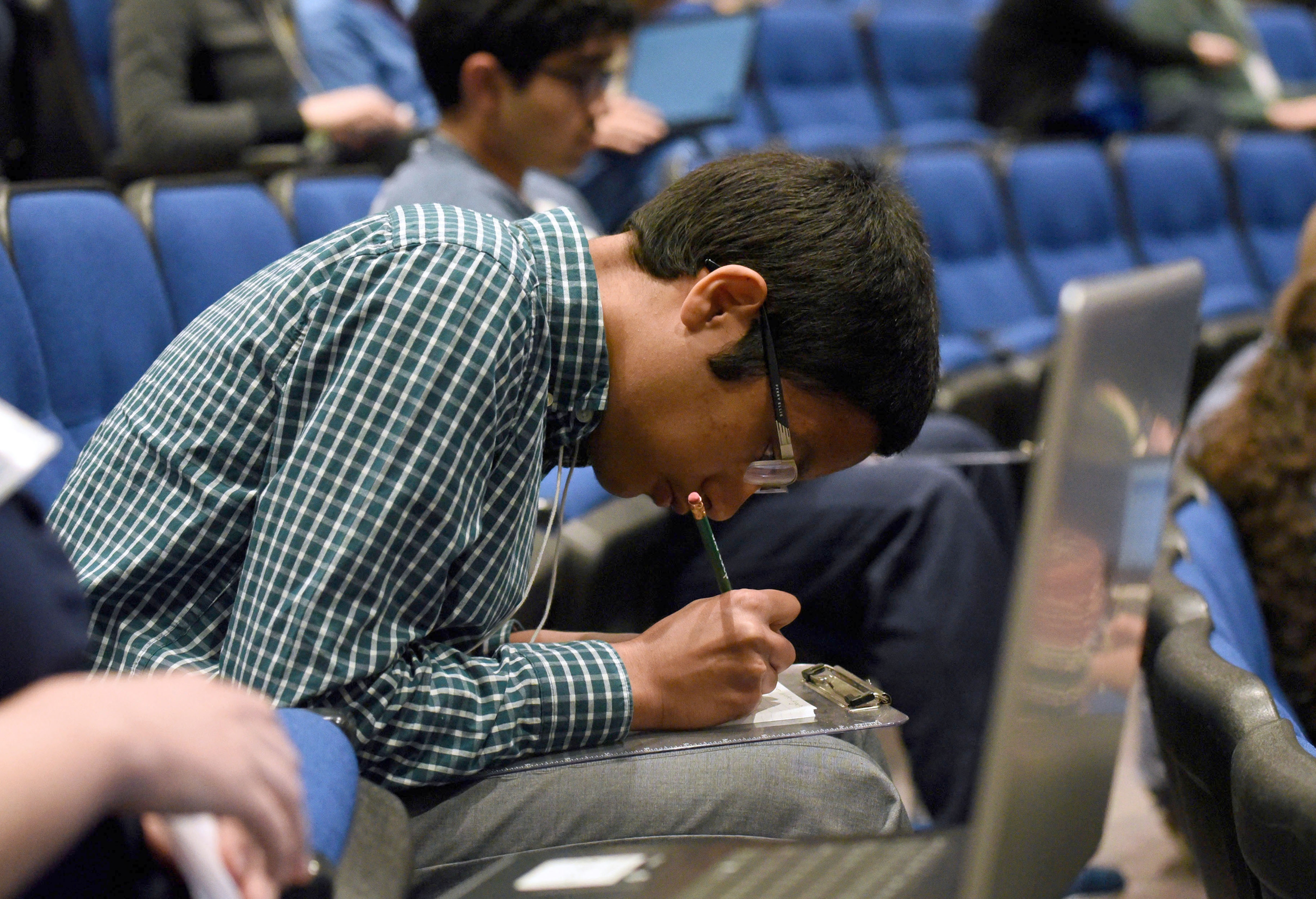 Cedar Cliff High School junior Thussentham Walter-Angelo, winner of the 2019 Central PA Regional Brain Bee, bends over a clipboard on his lap and writes answers during the 2019 USA National Brain Bee, held April 12-14 at Penn State College of Medicine. He is seated in an auditorium, surrounded by other students. He wears a plaid shirt, gray pants and glasses.