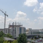 A large crane rises in to the blue sky and clouds above the glass, metal and cement of the Penn State Milton S. Hershey Medical Center complex. The buildings are surrounded by leafy trees and ever greens. Ribbons of blacktop form a circle around a grassy courtyard, the center of which contains a sculpture.