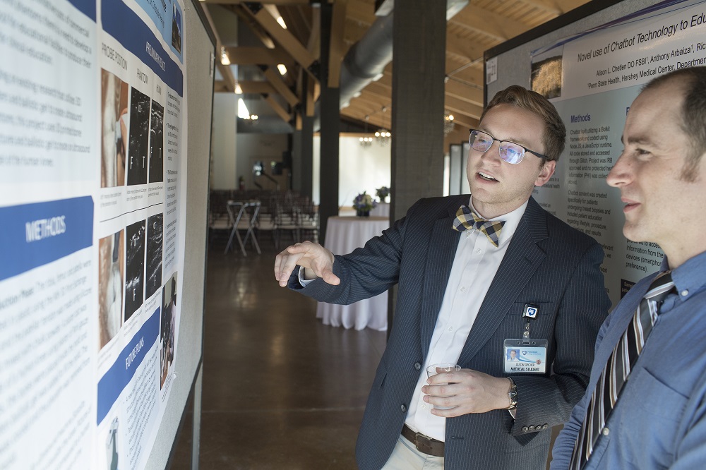 The poster at which Jason Spicher gestures is reflected in his glasses. He holds a glass in his other hand. Next to him, another man smiles at the poster. Behind them, support beams rise into the distance.