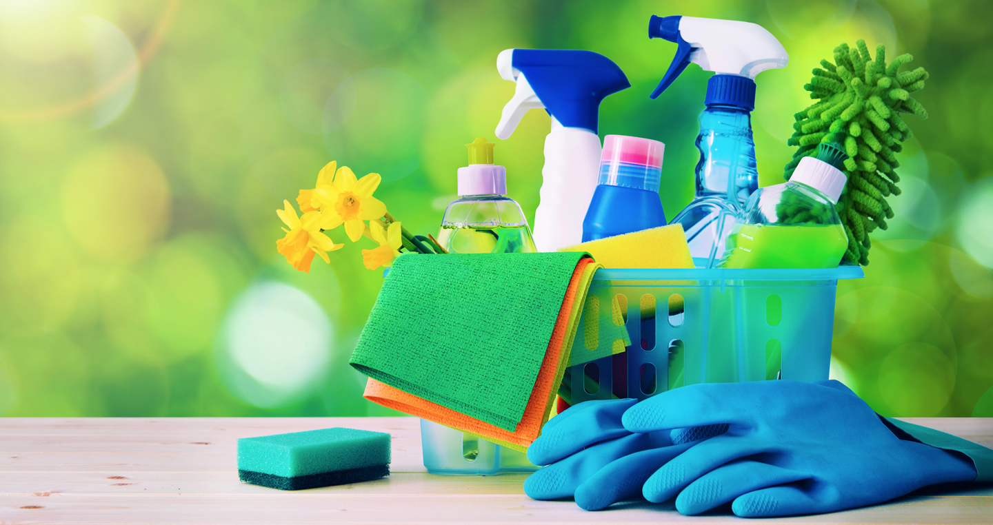 A plastic container with various cleaning supplies, including spray bottles and sponges, is on a table. Also sitting on the table is a pair of rubber gloves and a small sponge. Green foliage is in the background, out of focus.