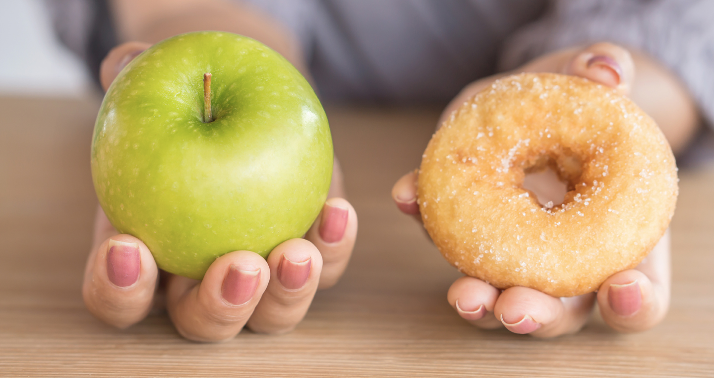 A close-up of two outstretched hands – one holding an apple tilted toward the camera, the other holding a doughnut tilted toward the camera.