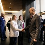 Gov. Tom Wolf, wearing a suit, shakes hands with a woman wearing a medical jacket. Other people in professional and medical attire look on. A hallway and large windows are in the background.