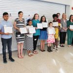 Fourteen people – 10 children and 4 adults – stand in a row to pose for a photo, all of them smiling. Each child holds an award certificate.
