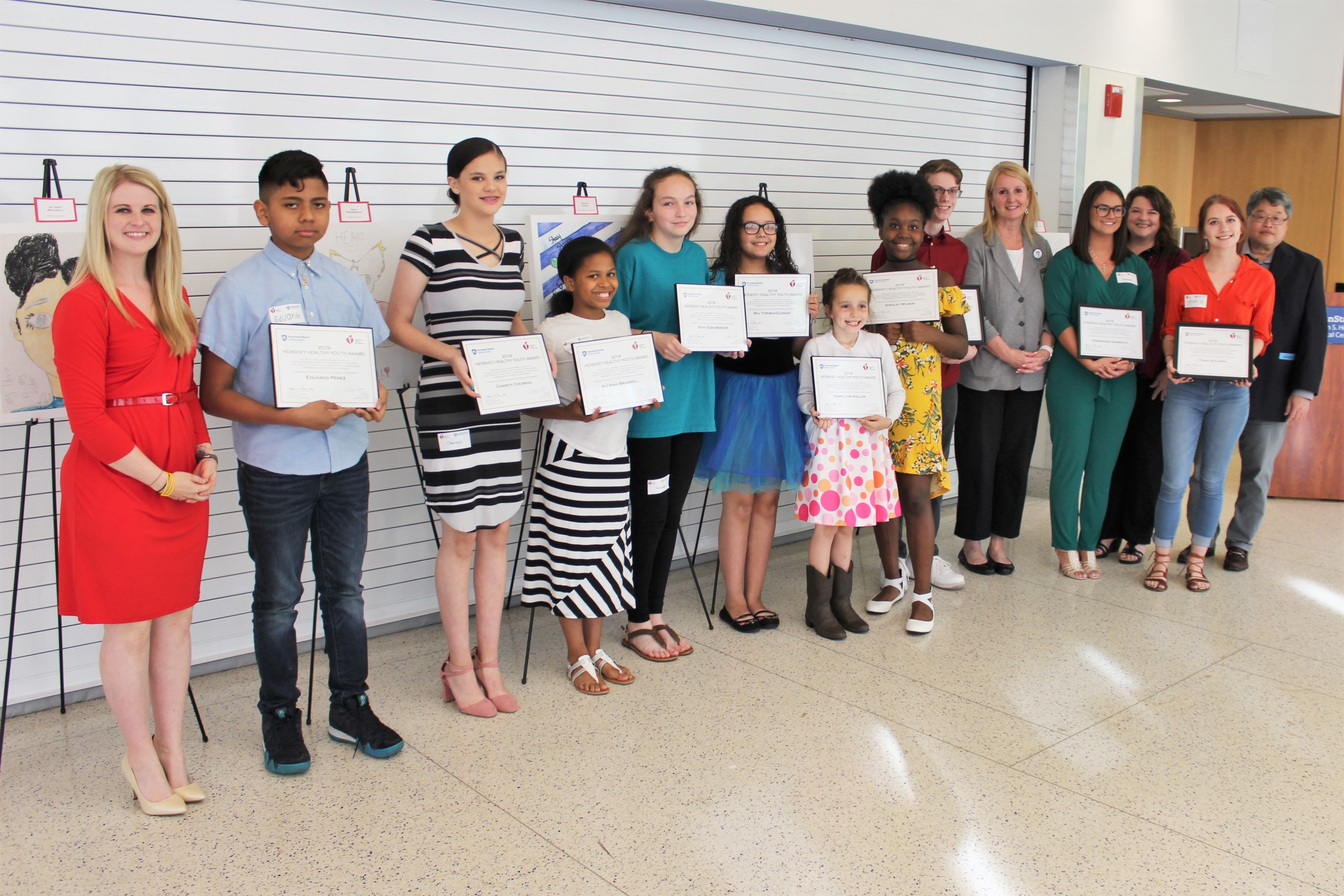 Fourteen people – 10 children and 4 adults – stand in a row to pose for a photo, all of them smiling. Each child holds an award certificate.