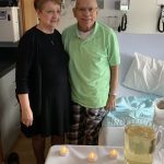 Bernadine and Charles Mineweaser stand in a hospital room with their arms around each other and smile. In front of them is a table with a cake, tea lights and a pitcher of sparkling grape juice. Next to Charles is a hospital bed, and equipment is behind them. Bernadine is wearing a dress, and Charles is wearing a polo shirt and plaid pajama pants.