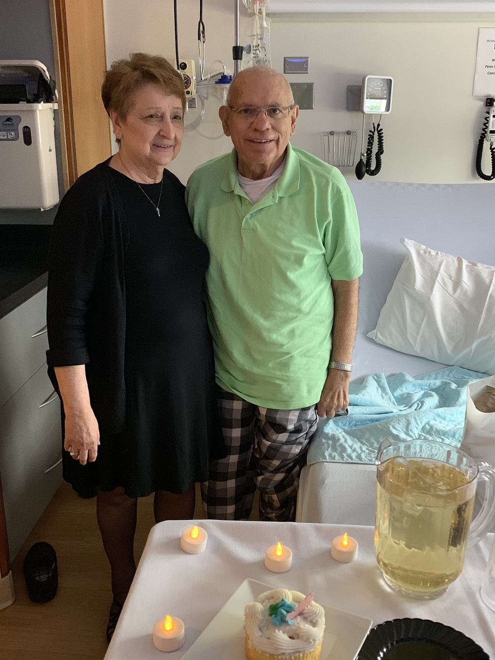 Bernadine and Charles Mineweaser stand in a hospital room with their arms around each other and smile. In front of them is a table with a cake, tea lights and a pitcher of sparkling grape juice. Next to Charles is a hospital bed, and equipment is behind them. Bernadine is wearing a dress, and Charles is wearing a polo shirt and plaid pajama pants.