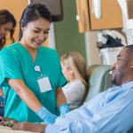 A woman in scrubs and rubber gloves holds a man’s arm which is blocked from view by his torso. The man squeezes a small rubber ball. Behind them, a woman has a similar interaction with another patient.