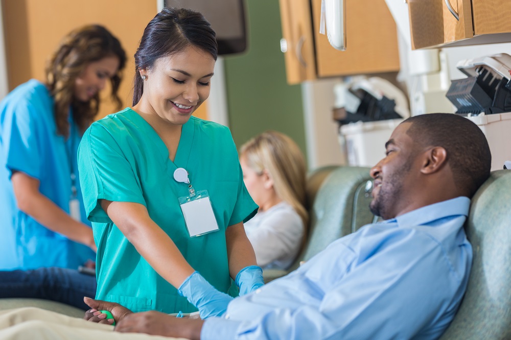 A woman in scrubs and rubber gloves holds a man’s arm which is blocked from view by his torso. The man squeezes a small rubber ball. Behind them, a woman has a similar interaction with another patient.