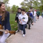 Marissa Aulenbach plans a guitar while glancing over her shoulder line of children making their way behind her down a brick path. Trees and green grass fill the frame behind them.
