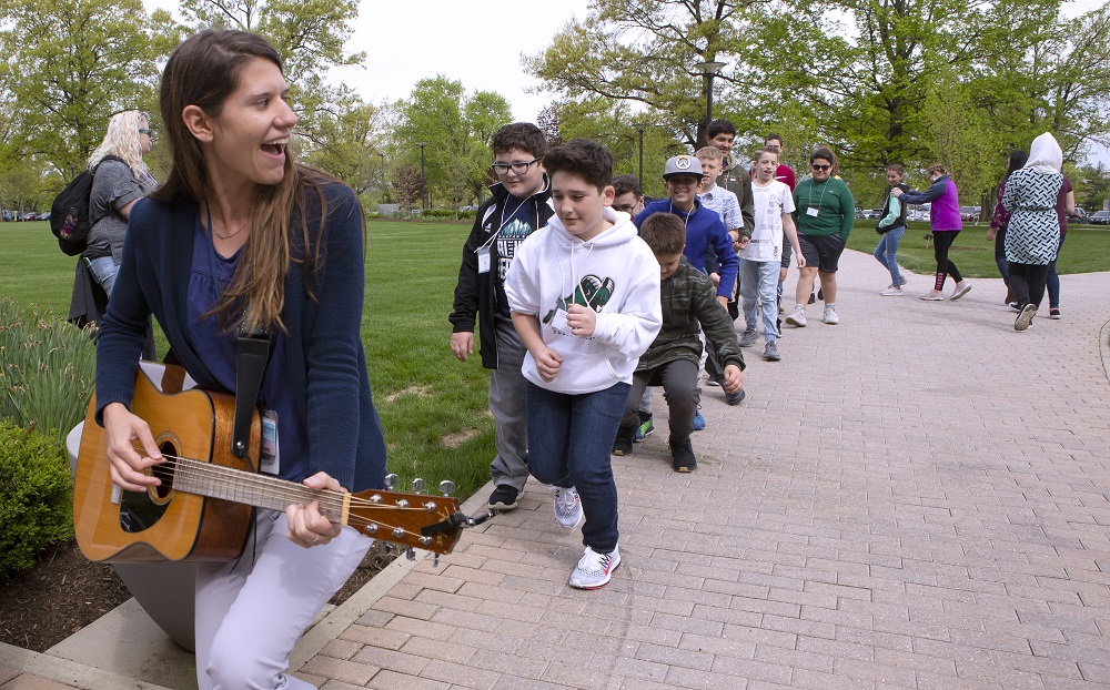 Marissa Aulenbach plans a guitar while glancing over her shoulder line of children making their way behind her down a brick path. Trees and green grass fill the frame behind them.