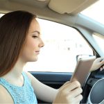 Side view of a teenage girl as she drives. Her left hand is on the steering wheel. Her right hand holds up a cell phone, and she’s looking to the right at the phone instead of looking at the road.