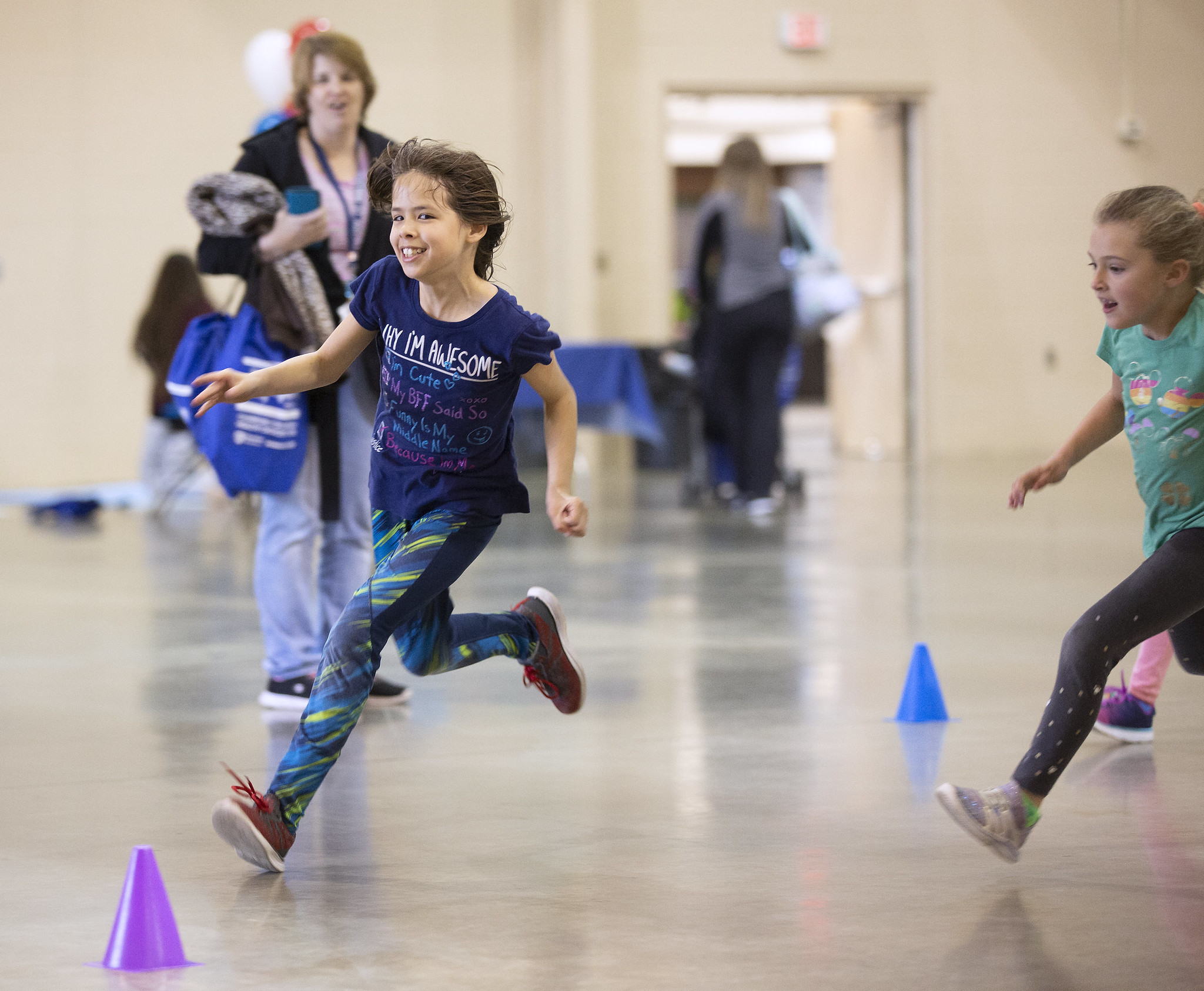 A young girl dressed in a t-shirt, long pants and sneakers smiles as she runs around a cone set up in a gymnasium. Another girl is close behind her, as a woman looks on in the background.