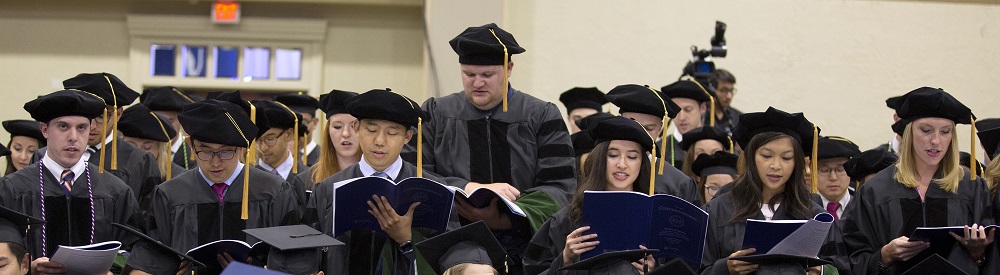 A row of graduates in traditional caps and gowns read from folders full of papers.