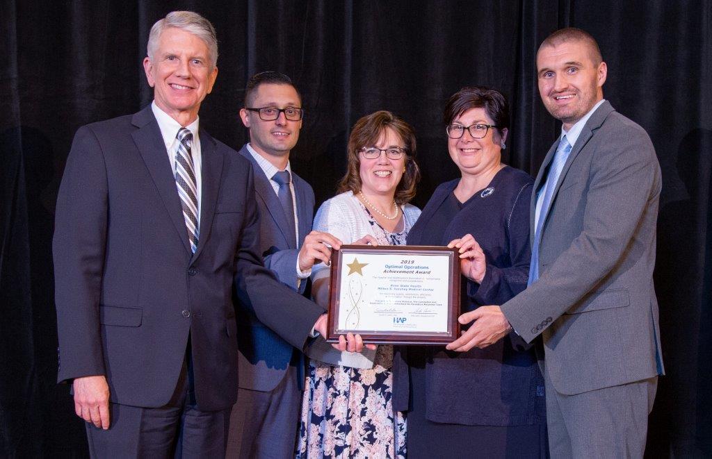 Five people – two women and three men, pose for a photo, wearing semi-formal/business attire, each with a hand on a framed certificate that reads “2019 Optimal Operations Achievement Award.”