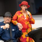 Al Dolatoski and his wife Joyce hold hands and hold microphones as they sing Sonny and Cher’s “I Got You Babe” during the LVAD Celebration of Life. Al is sitting in a wheelchair and wearing a sweatshirt and hat. He has an LVAD hanging by a strap around his shoulder. Joyce is wearing a flowered dress and hat.
