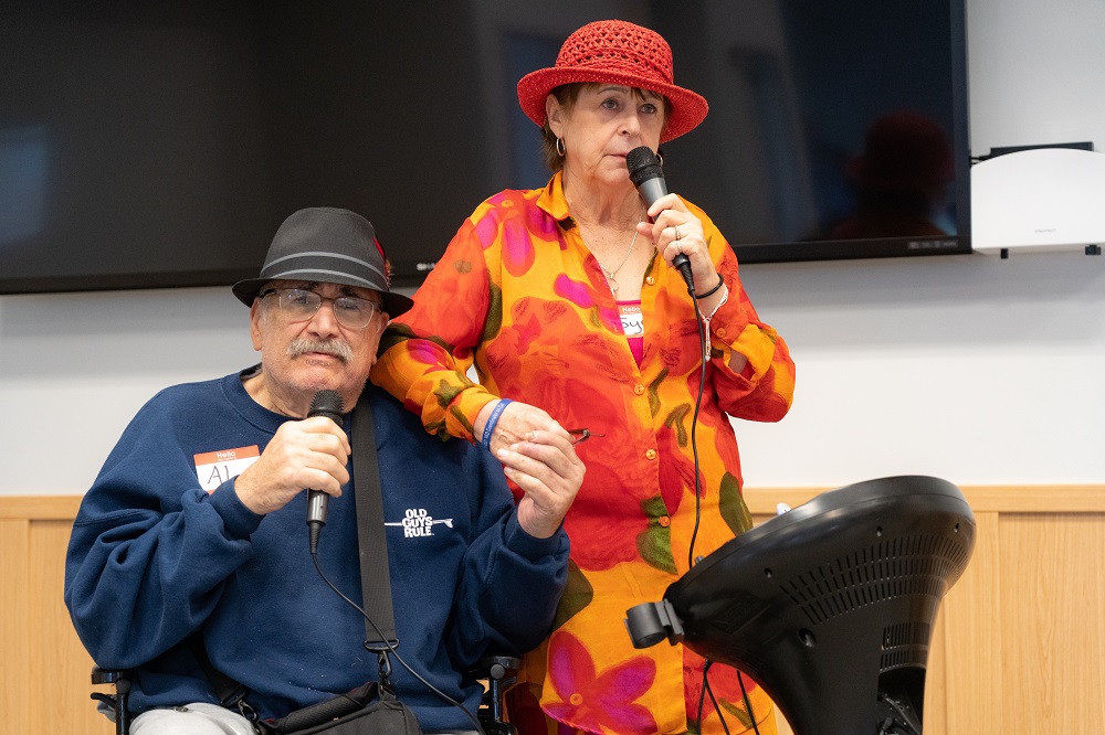 Al Dolatoski and his wife Joyce hold hands and hold microphones as they sing Sonny and Cher’s “I Got You Babe” during the LVAD Celebration of Life. Al is sitting in a wheelchair and wearing a sweatshirt and hat. He has an LVAD hanging by a strap around his shoulder. Joyce is wearing a flowered dress and hat.
