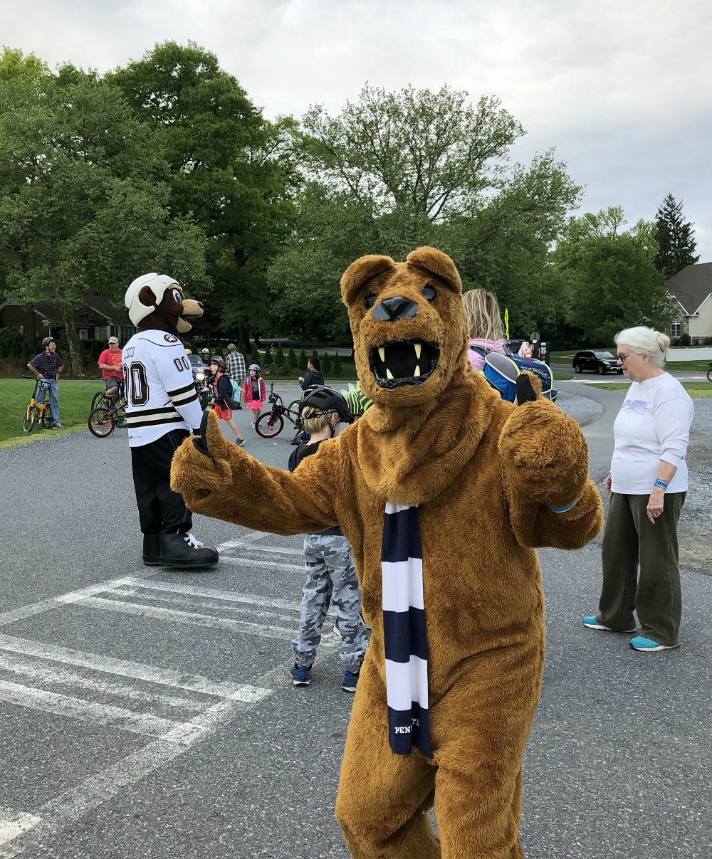 Someone in a costume dressed as the Penn State Nittany Lion gives a thumbs up sign. Behind him people on bicycles gather in a parking lot. A man dressed as the Hershey Bear’s mascot, Cocoa, stands among them.