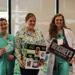 Three women, two in scrubs, smile and hold prizes – tickets, a blender and a large Hershey’s Milk Chocolate bar.