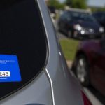 A Penn State Milton S. Hershey Medical Center parking permit sticker is on the bottom-right corner of a vehicle’s rear window. Other cars are seen out-of-focus in the parking lot.