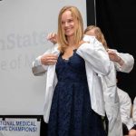 Kerri Schopf, wearing a dress, smiles as another woman stands behind her and slips a white coat onto her shoulders. Behind her, the words Penn State College of Medicine are projected onto a screen.