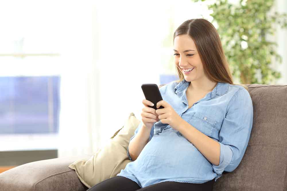 A woman smiles at her cellphone while reclining on a couch. Behind her, out of focus, are a potted plant and a window.