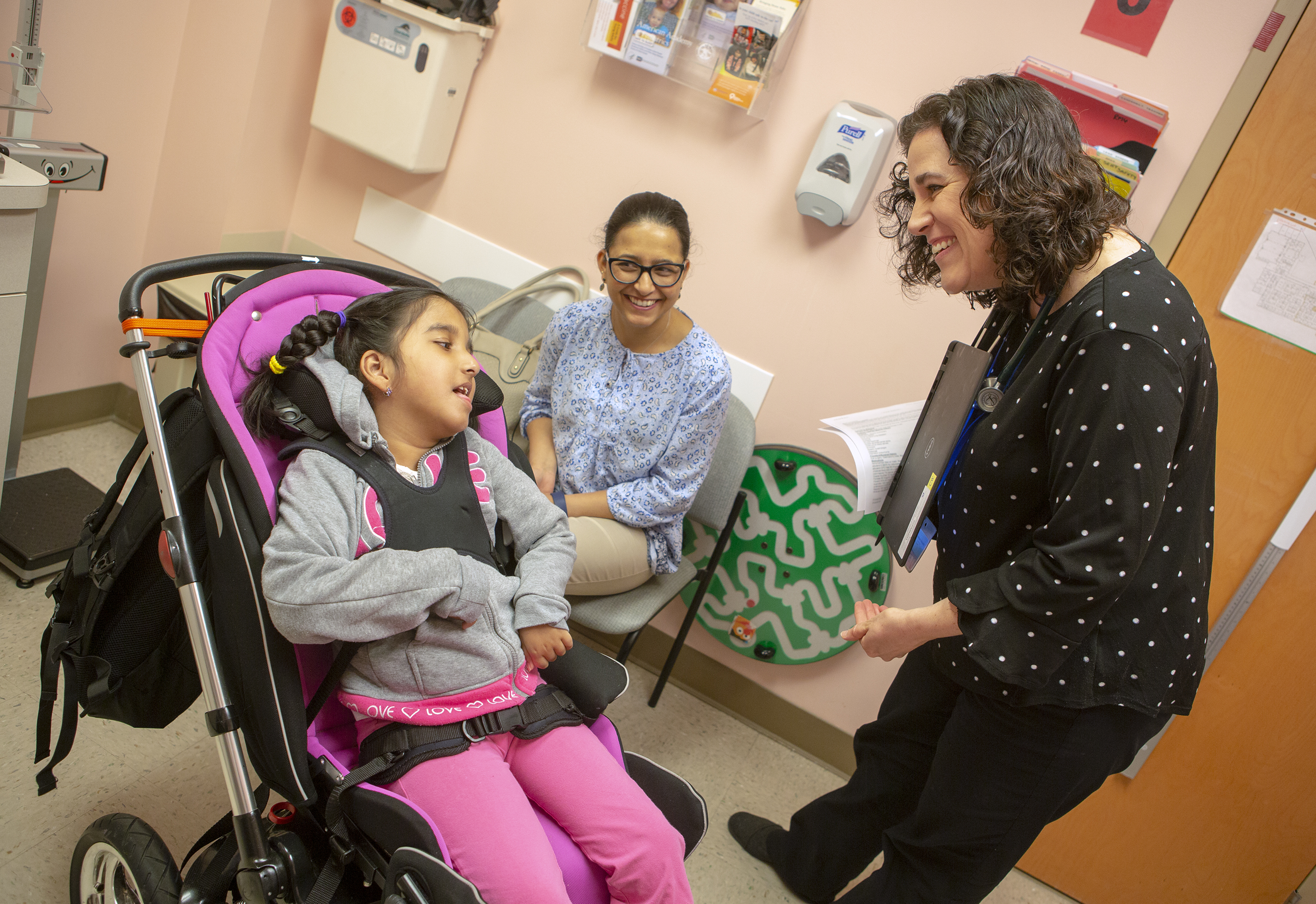 Dr. Laura Murphy, pediatrician with Penn State Children’s Hospital’s Pediatric Complex Care Program, smiles and bends down to greet her patient, Brinda Rizal, who is in a wheelchair. Brinda, who has braids and is wearing a sweatshirt and pants, is strapped into the wheelchair and looks up at Murphy, who is wearing a polka dotted shirt and pants and wears a stethoscope around her neck. Brinda’s mother, Basudha Rizal, wearing glasses and a printed top and pants, is sitting in a chair against the wall. She smiles at her daughter. A soap dispenser, pamphlet rack and folders are hanging on the wall of the exam rom.