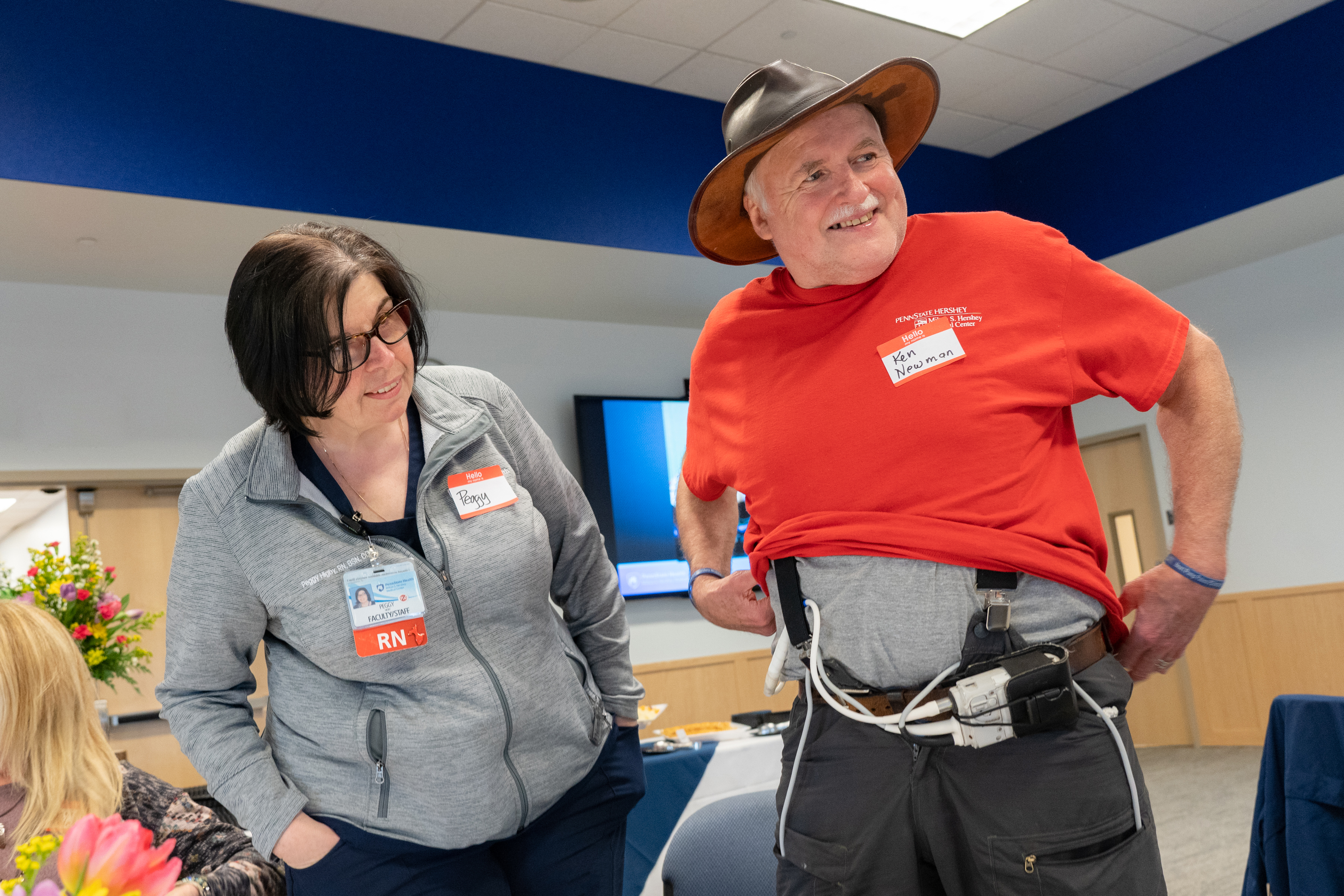 Ken Newman, right, holds up his shirt to show a HeartMate II LVAD attached to his belt. He is wearing a hat, a nametag and has a gray moustache. Peggy Higby, the LVAD and heart transplant manager, stands next to him. She is wearing glasses and is wearing a sweat jacket with a nametag and an RN badge.