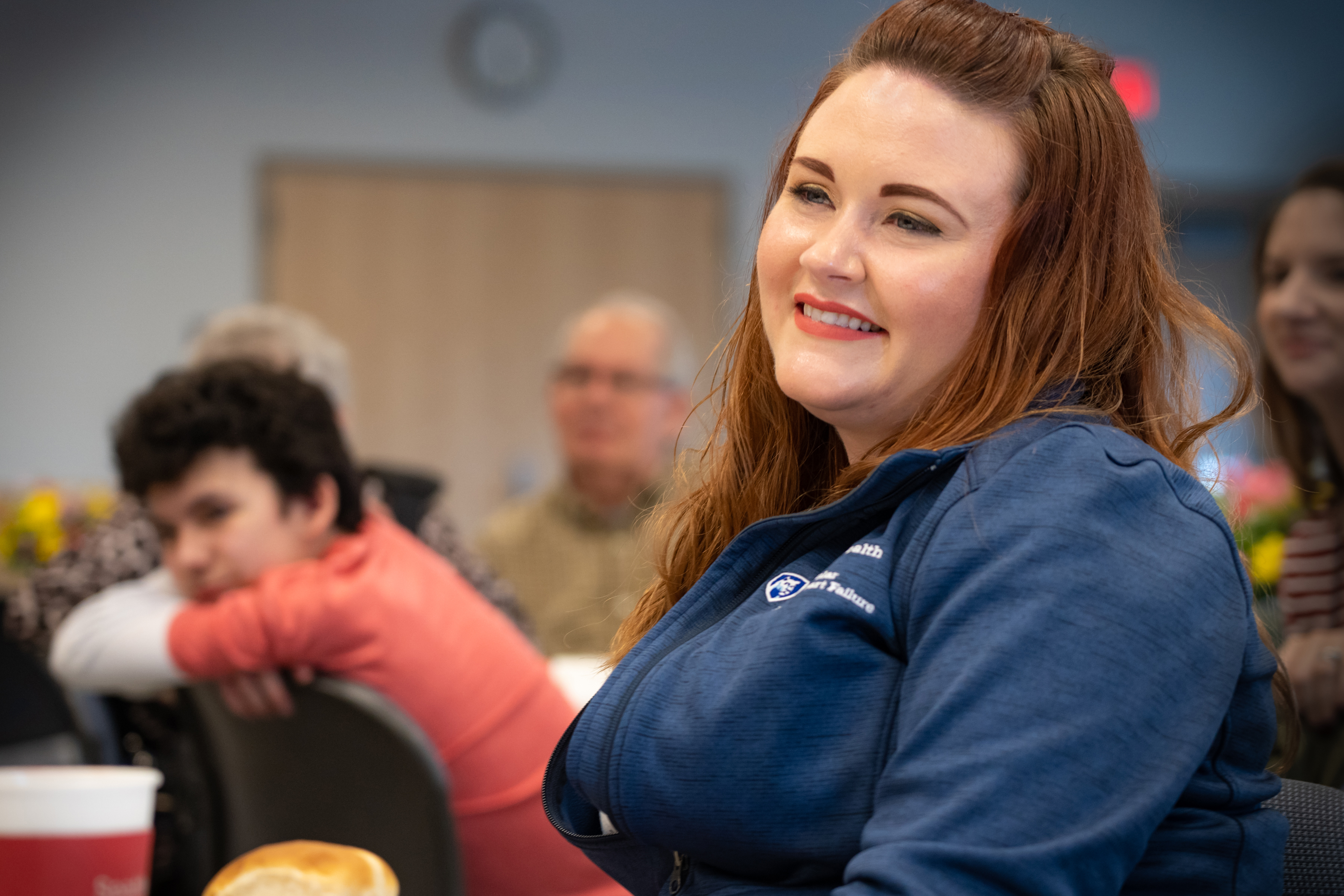 Transplant assistant Aimee Zehring smiles as she listens to Al Dolatoski’s thank you speech during the LVAD reunion. She has long hair and is wearing a Penn State Health jacket. Behind her several people sit at tables, shown out of focus.