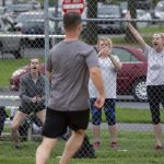 Through a chain link fence, six women cheer for a man in shorts and a T-shirt who is running past them.