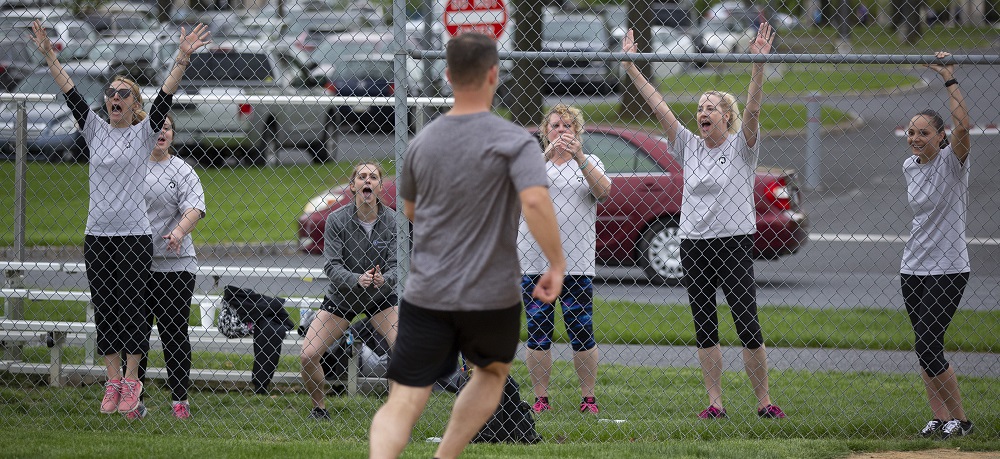 Through a chain link fence, six women cheer for a man in shorts and a T-shirt who is running past them.